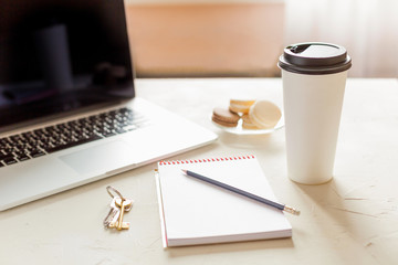 Hot coffee in a paper cup, notebook and a laptop  on the white table. Business breakfast. 