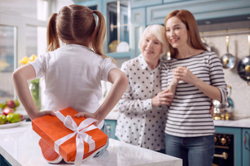 Pleasant surprise. Charming little girl sitting on the table and hiding a gift box behind her box,...