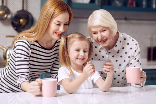 Dearest people. Beautiful little girl sitting at the kitchen counter and taking a selfie with her beloved mother and grandmother while the women holding cups of coffee