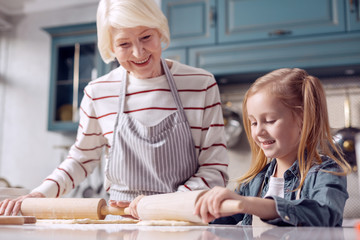 Cooking lessons. Charming little girl standing next to her grandmother and rolling out dough...