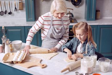 Sweet tooth. Adorable little girl helping her grandmother to make cookies, cutting out different figures, while the woman rolling out some more dough