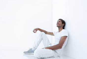 dreamy young man sitting beside a white wall.