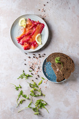 Sliced beetroot marinated salmon for sandwiches with sliced rye bread, pink salt, pepper, greens and lemon served in blue plates over grey texture background. Top view, space