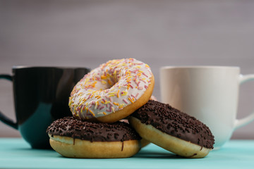 Three donuts, one on top of another, chocolate and white cream. Behind them are two mugs with a hot drink.