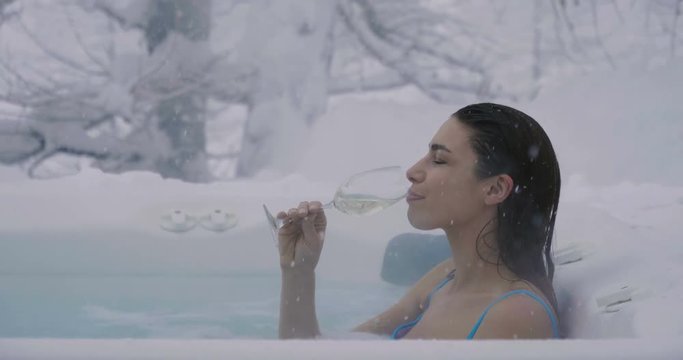 A beautiful woman drinks champagne in a hot tub surrounded by nature as the snow falls from the sky. Concept of: relaxation, nature, wellness and vacation.
