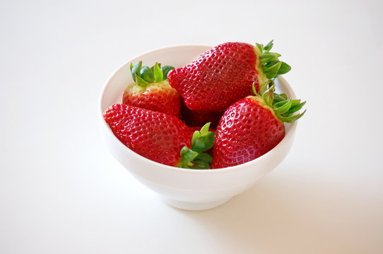 Strawberries lie in a white bowl on a white surface.