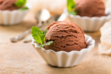 Chocolate ice cream scoop in white bowl with mint leaves on sand color stone background. Summer food concept with copy space.