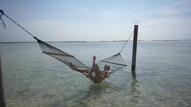 Female tourist relaxes in a hammock above shallow ocean waters on a sunny day