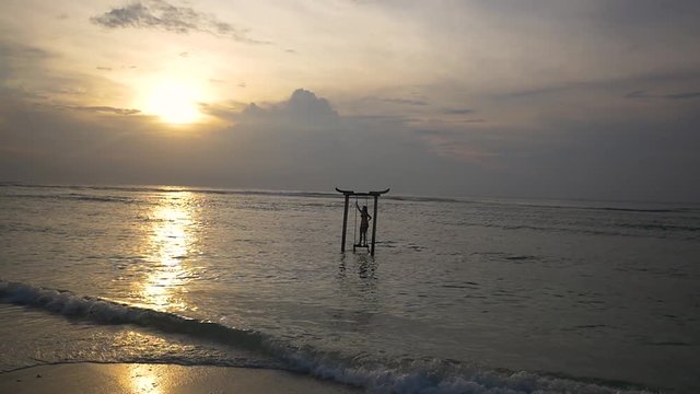 Silhouette of woman standing on wooden swing in the ocean under beautiful sunset