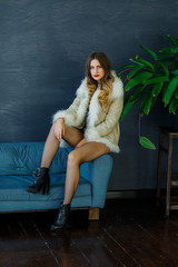 A beautiful sexy blonde in a light sheepskin coat sits on the couch and poses in the interior. Fashion, beauty. Winter collection.