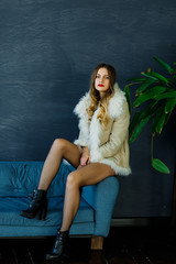 A beautiful sexy blonde in a light sheepskin coat sits on the couch and poses in the interior. Fashion, beauty. Winter collection.