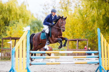 Young rider man on bay horse jumping over hurdle on show jumping competition