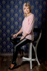 Portrait of a beautiful blonde woman in a pink jacket and leather pants in a classic interior with blue wallpaper with monograms