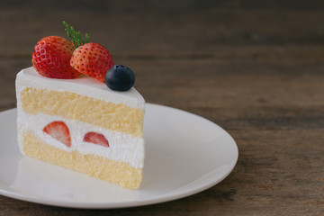 Strawberry shortcake on white plate put on wood table with copy space. Delicious and soft vanilla sponge cake with dairy whipped cream and fresh strawberries and blueberry. Homemade bakery concept.