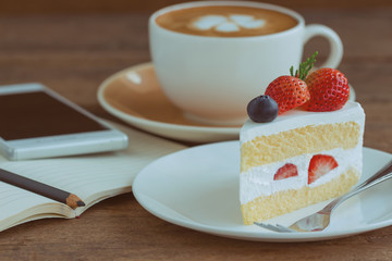 Homemade delicious and soft strawberry shortcake on wood table beside smartphone and hot cappuccino coffee at bakery shop or cafe restaurant. Vanilla sponge cake with whipped cream and strawberries.