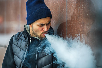Vape man. Portrait of a handsome young white guy in black waistcoat and modern cap vaping an...