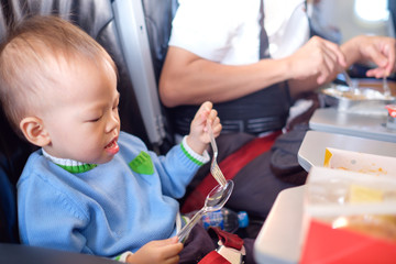 Cute little Asian 18 months / 1 year old toddler baby boy child wearing blue sweater eating food during flight on airplane. Flying with children, Happy air travel with kids & little traveler concept