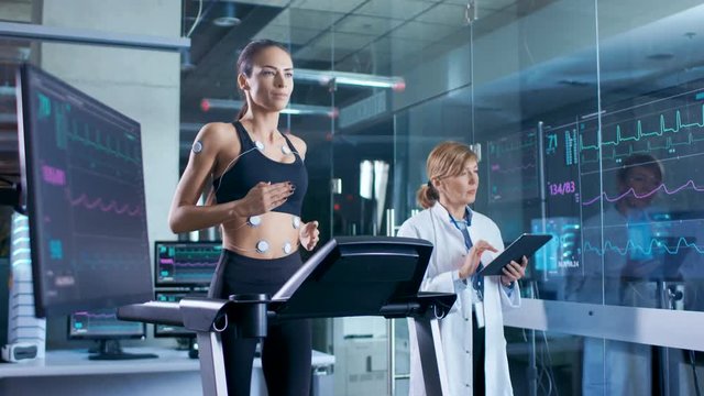 Beautiful Woman Athlete Runs on a Treadmill with Electrodes Attached to Her Body, Female Physician Uses Tablet Computer and Controls EKG Data Showing on Laboratory Monitors.