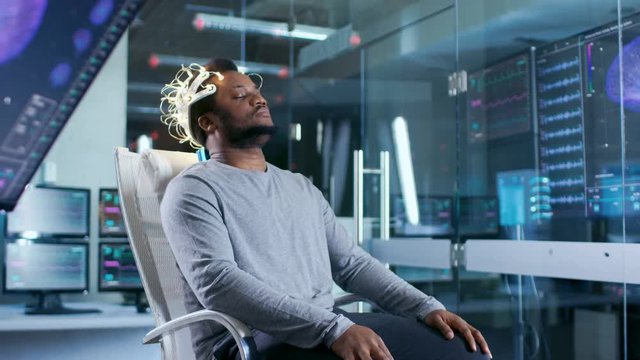 In Laboratory Man Wearing Brainwave Scanning Headset Sits in a Chair with Closed Eyes. In the Modern Brain Study/ Neurological Research Laboratory. Monitors Show EEG Reading and Graphical Brain Model.