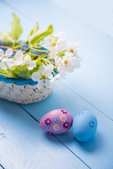 Obraz na płótnie Canvas Two painted Easter eggs near basket with white spring flowering branch on light blue background