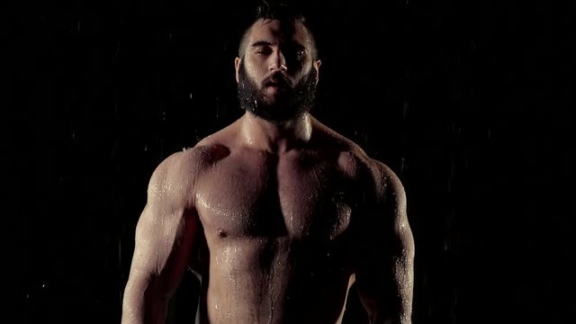 wet exhausted muscular bodybuilder with naked torso is standing in dark room and looking in camera