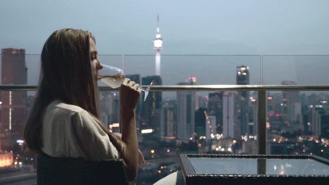 Happy woman drinking a glass of white wine while sitting in rooft bar in the city or on the terrace with cityscape at sunset. Vacation,happiness,freedom concept.