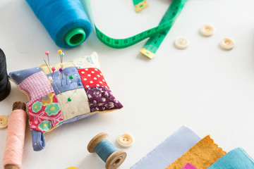 Obraz na płótnie Canvas needlework, craft, sewing and tailoring concept - tools closeup on white desk, measuring meter, blue, pink and black thread spools , scraps of colorful fabric, pins and pincushion, white buttons.