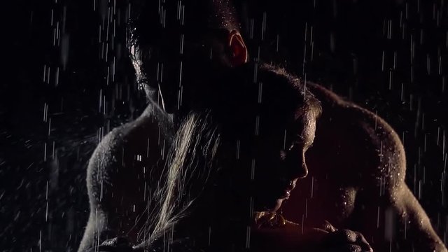 couple of lovers is standing under rain in darkness, brawny man is hugging her woman shoulders