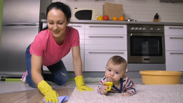 Happy cheerful hispanic mother and housewife scrubbing kitchen floor in rubber gloves at home. Adorable baby son crawling on carpet. Sweet family doing housework together. Slow motion. Dolly shot