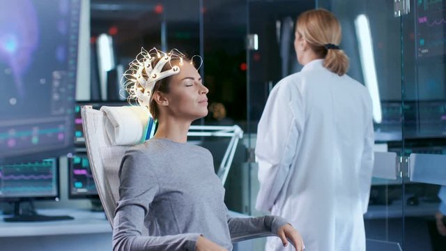 Woman Wearing Brainwave Scanning Headset Sits in a Chair while Scientist Supervises. In the Modern Brain Study Laboratory Monitors Show EEG Reading and Brain Model. 