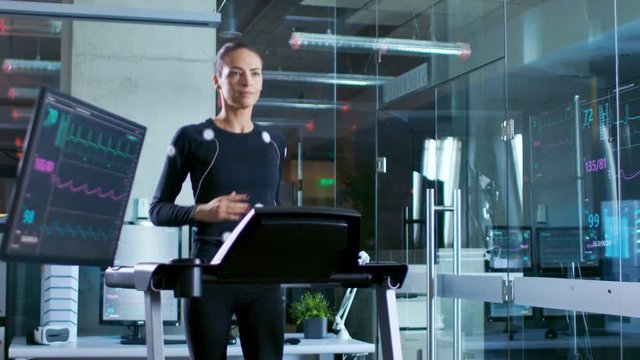 In Scientific Sports Laboratory Beautiful Woman Athlete Runs on a Treadmill with Electrodes Attached to Her Body, Monitors Show EKG Data on Display. Slow Motion.