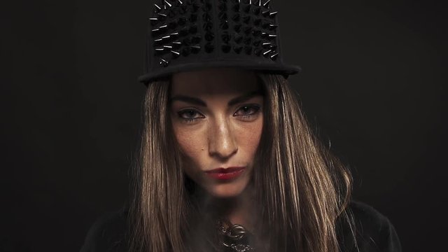 Young pretty caucasian brunette woman with red lipstick in spiked snapback cap blows vape smoke out nose on black background