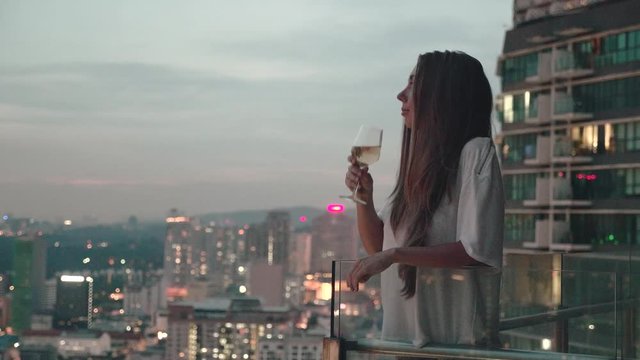 Happy woman drinking a glass of white wine while sitting in rooft bar in the city or on the terrace with cityscape at sunset. Vacation,happiness,freedom concept.