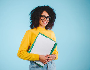Obraz premium Portrait smiling young african woman in glasses holding folder over blue background. Education concept.