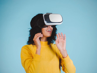 Obraz premium Future is right now. Confident young african american woman adjusting her virtual reality headset and smiling on blue background. Girl in VR goggles