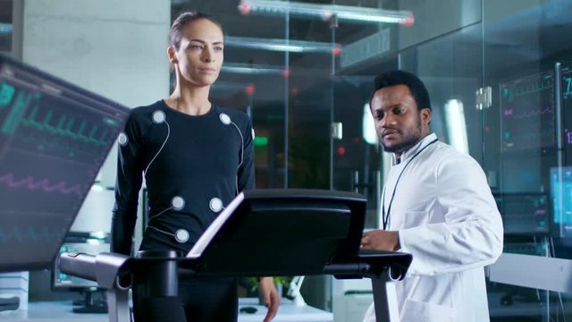 In Scientific Sports Laboratory Beautiful Woman Athlete Walks on a Treadmill with Electrodes Attached to Her Body, Monitors Show EKG Data on Display. Slow Motion.