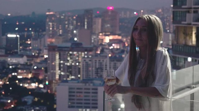 Happy woman drinking a glass of white wine while sitting in rooft bar in the city or on the terrace with cityscape at sunset. Vacation,happiness,freedom concept.