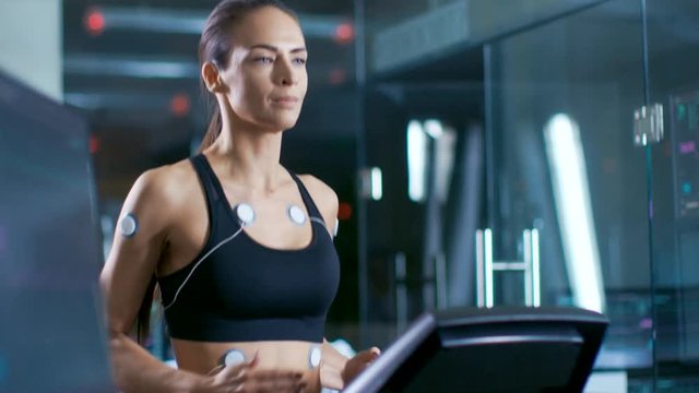 In Scientific Sports Laboratory Beautiful Woman Athlete Runs on a Treadmill with Electrodes Attached to Her Body, Monitors Show EKG Data on Display. Slow Motion.