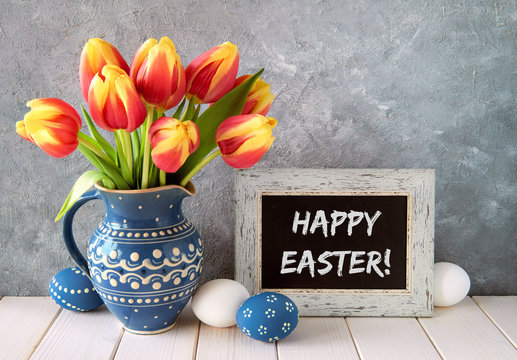 Red-yellow tulips in blue ceramic pitcher with Easter eggs and a blackboard, text