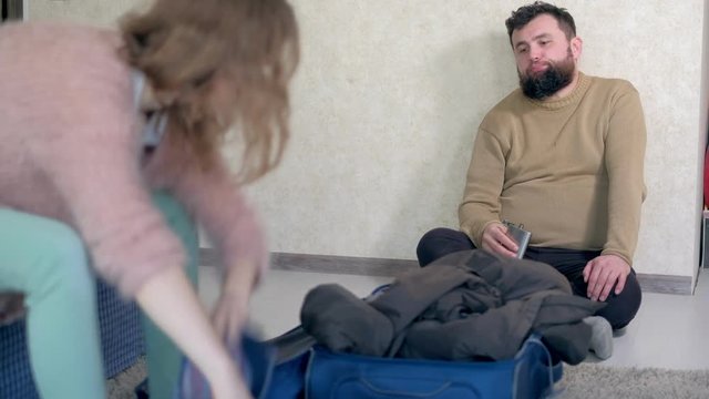 Disappointed,crying and beaten young woman packing her clothing in the suitcase, drunken, drinks from a metal flask,he takes out and throws her stuff from the bag,conceptual scene.
