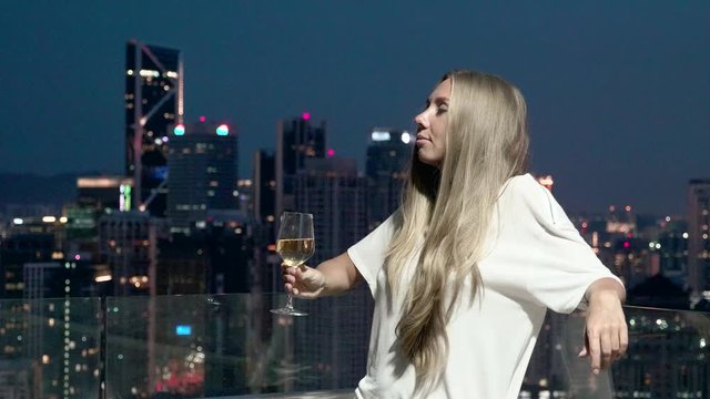 Happy woman drinking a glass of white wine while sitting in rooft bar in the city or on the terrace with cityscape at sunset. Vacation,happiness,freedom concept.