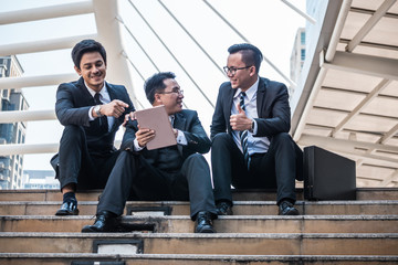 Happy Asian business men working outdoor with tablet, Group of people talking together while sitting on stairs in front of the office building.