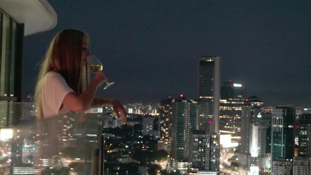 Happy woman drinking a glass of white wine while sitting in rooft bar in the city or on the terrace with cityscape at sunset. Vacation,happiness,freedom concept.