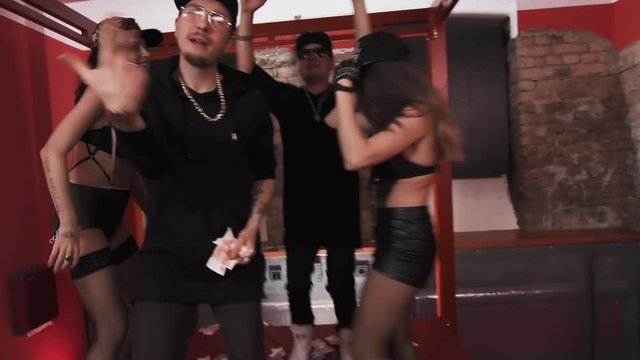 Cheeky caucasian rappers in black clothes glasses and snapbacks with sexy girls in underwear standing on bed covered in money rapping