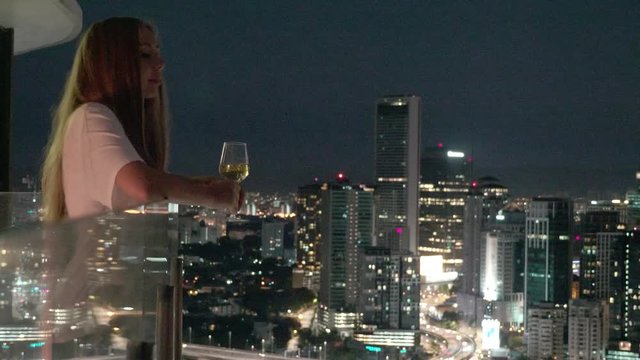 Happy woman drinking a glass of white wine while sitting in rooft bar in the city or on the terrace with cityscape at sunset. Vacation,happiness,freedom concept.