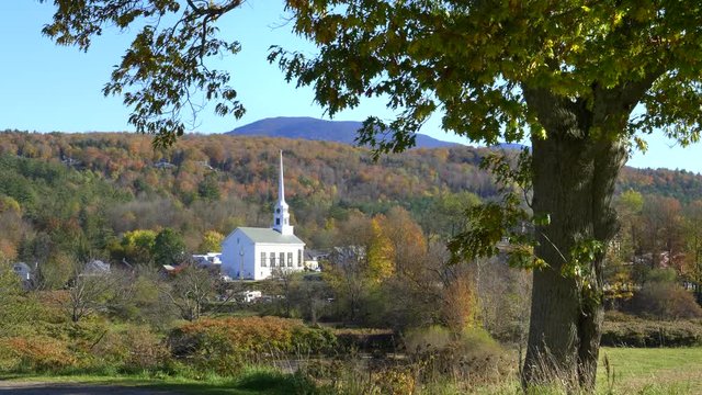 a white church at stowe framed by a tree and a hill with fall foliage in vermont, usa