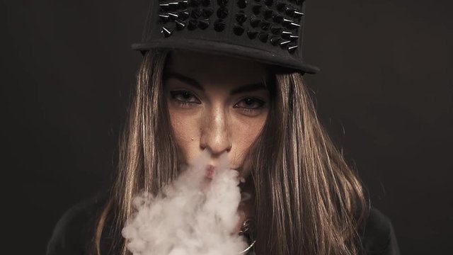 Young attractive caucasian brunette girl with red lipstick in spiked snapback cap blows vape smoke out nose on black background