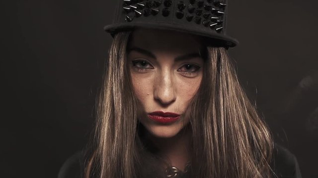 Young attractive caucasian brunette woman with red lipstick in spiked snapback cap blows vape smoke out nose on black background
