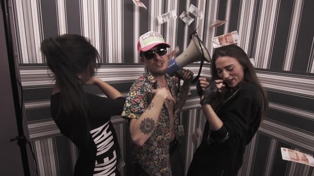 Impudent caucasian tattooed guy in sunglasses with sexy girls in black dresses dancing and rapping into megaphone, throw money, at apartment, striped wallpaper