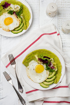 Spinach green crepes (pancakes) with fried egg, avocado and leaves of mix of salad on ceramic plate on white wooden background. Сoncept of healthy breakfast. Selective focus. Top view. Copt space.